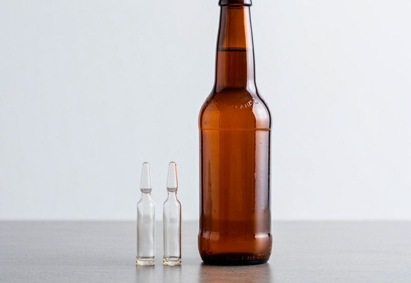 Закрытая коричневая бутылка пива стоит рядом с двумя стеклянными ампулами на серой поверхности